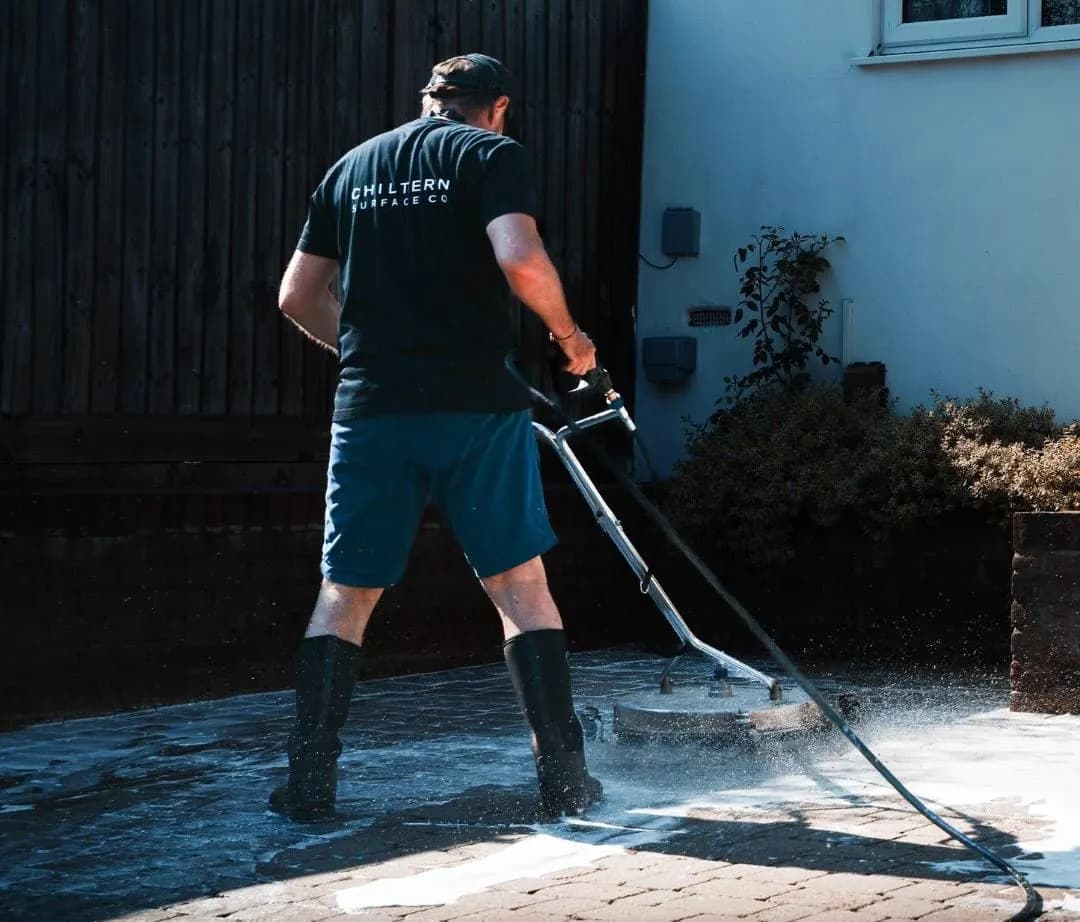Operative guiding a rotary surface cleaner across foamed block paving beside a rendered wall and garden shrubs