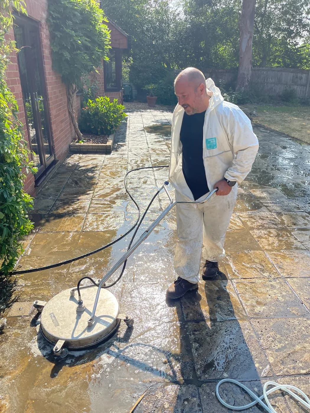 Operative in white overalls steering a rotary surface cleaner across a wet riven sandstone patio beside a brick house