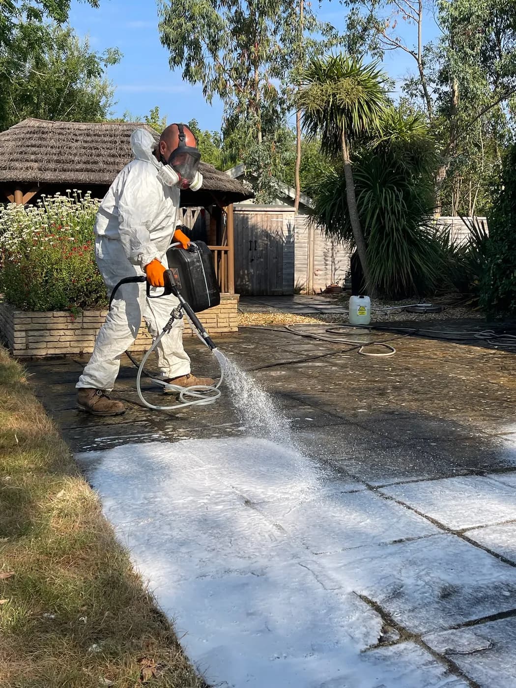 Operative in a white coverall and respirator spraying a foaming treatment across stone-effect patio slabs in a planted garden