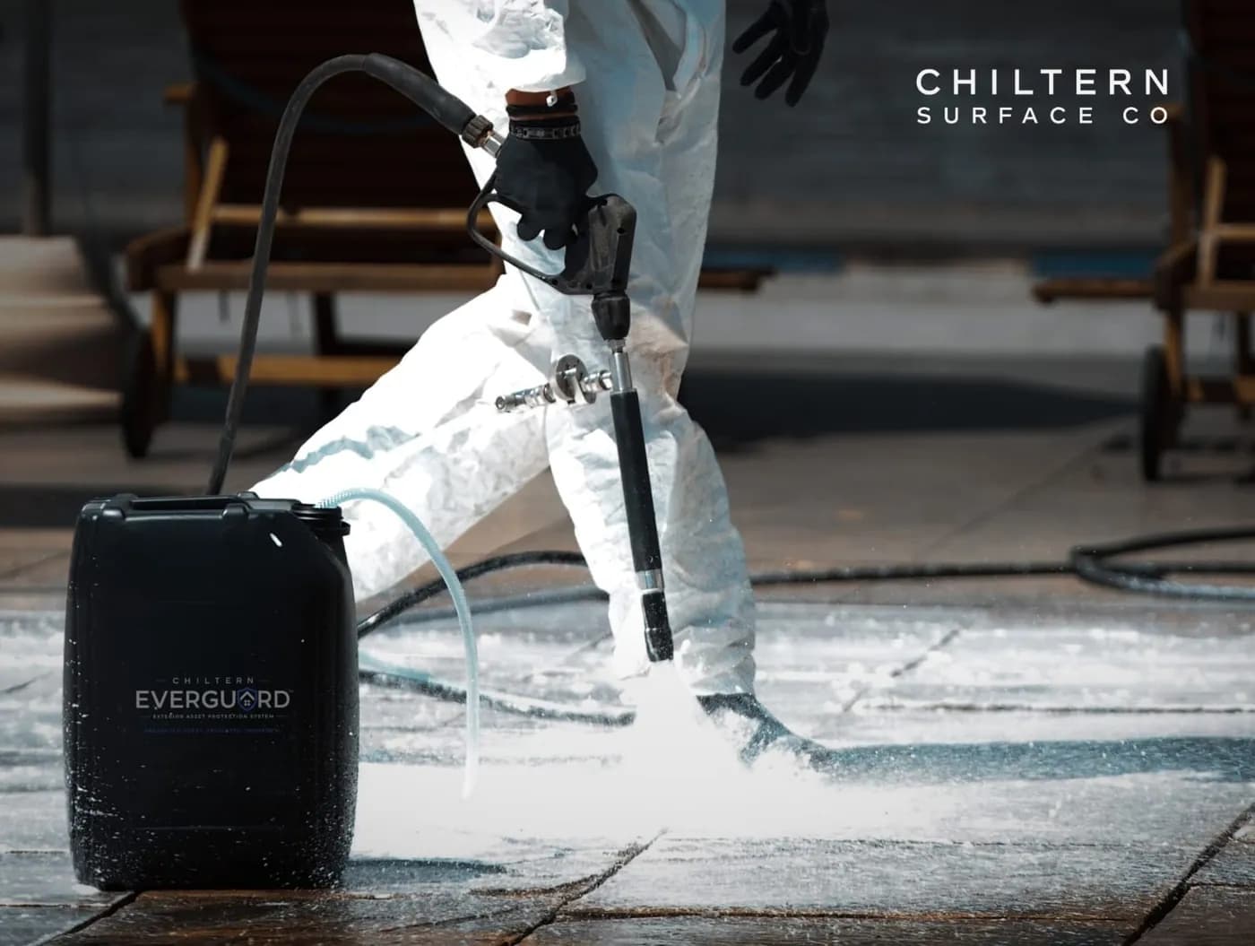 Operative in a white protective suit spraying a foaming treatment over stone paving beside a black Chiltern EverGuard™ drum