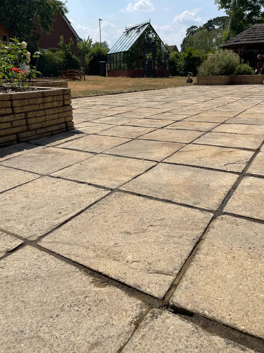 Low view across cleaned buff stone-effect patio slabs with sanded joints, a raised planter and a glasshouse beyond