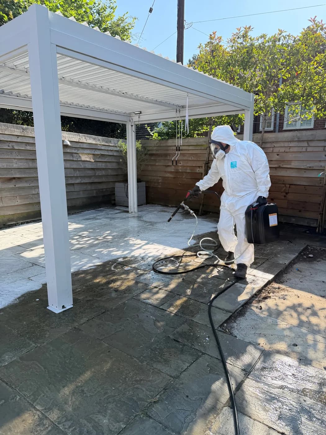Operative in a protective suit and full-face mask applying treatment to sandstone paving beneath a white pergola