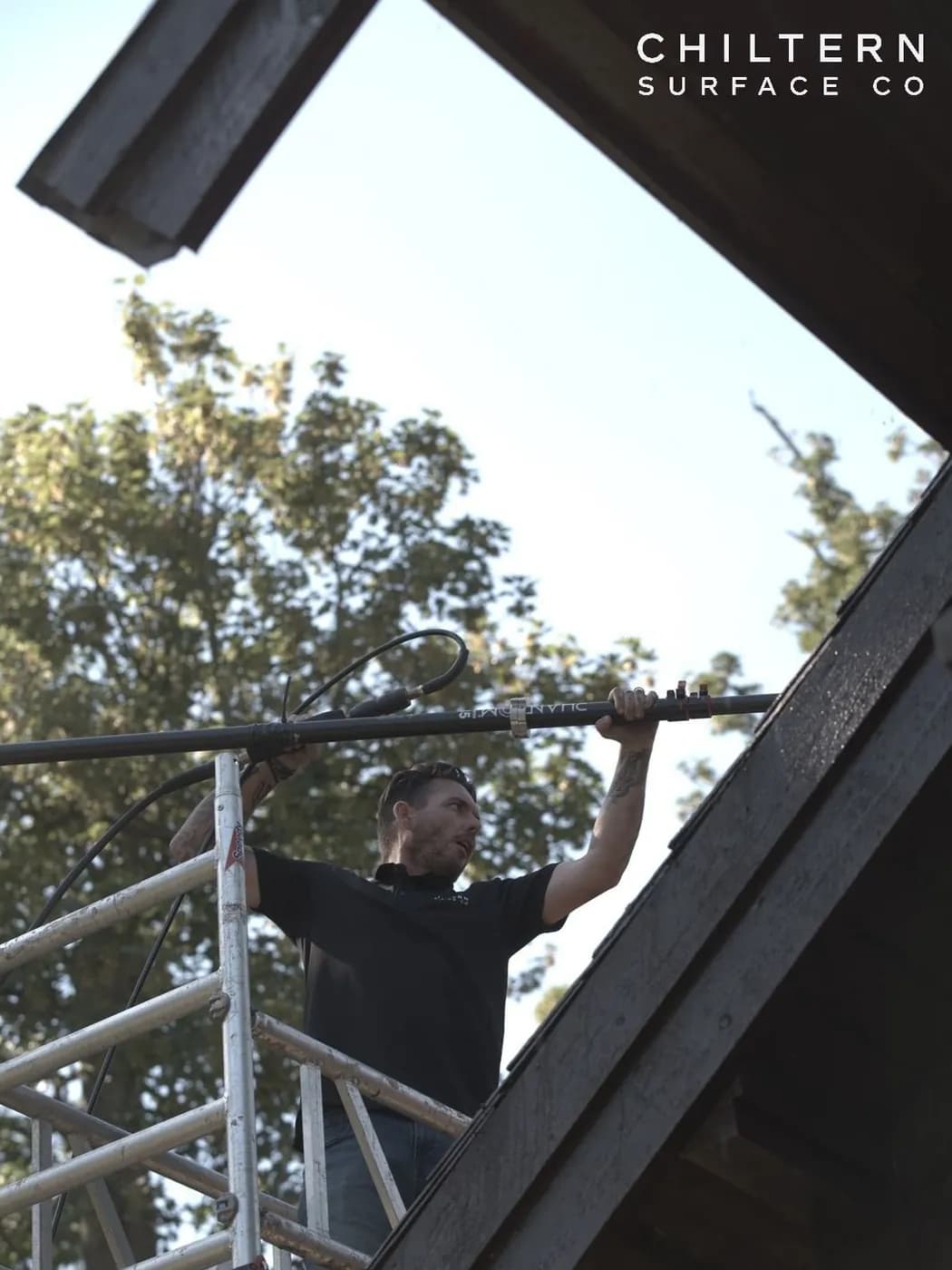 Operative on an aluminium access tower guiding a long pole along the edge of a roof, with a tall tree behind him