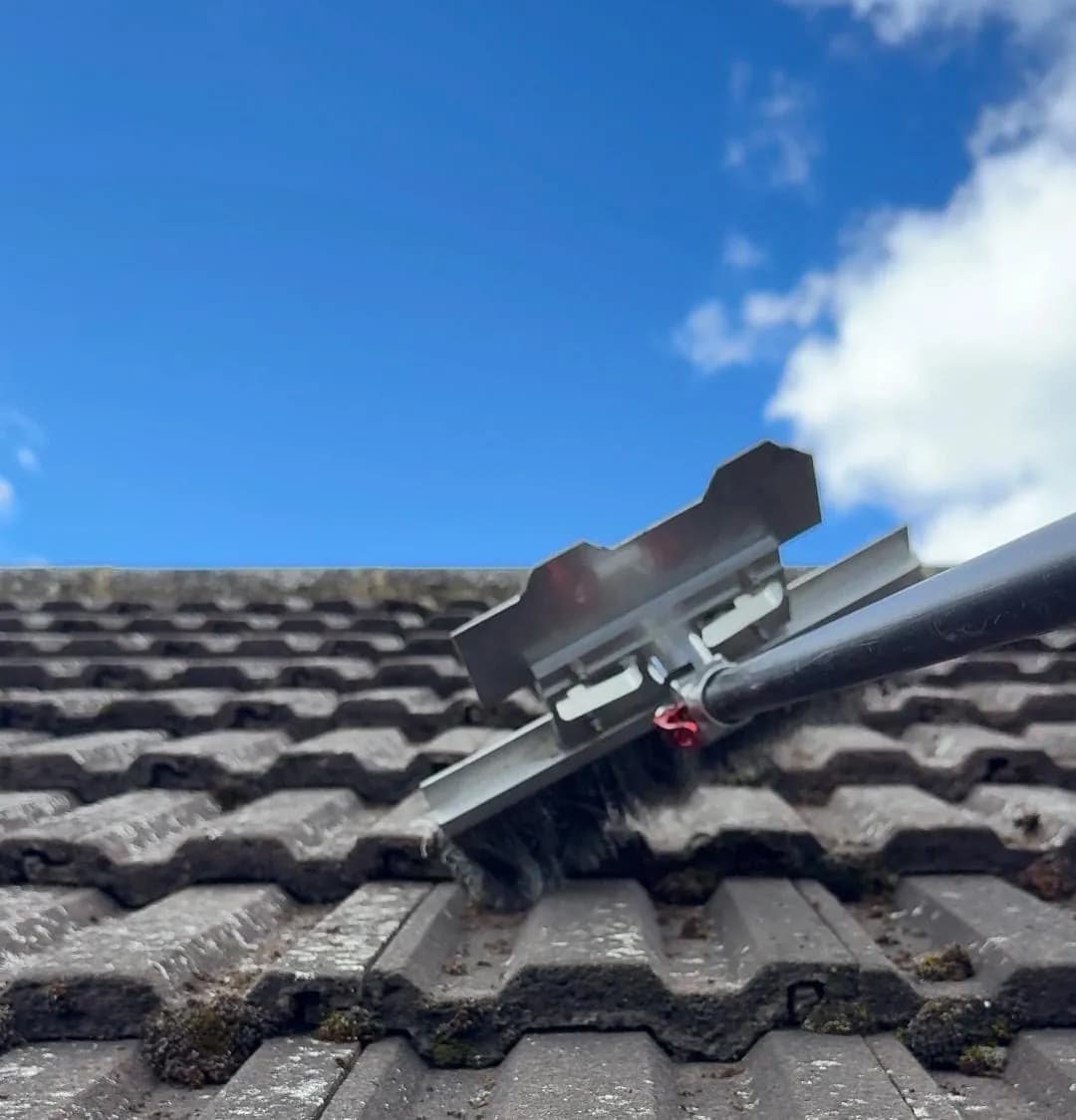 Pole-mounted scraper and brush lifting moss from the laps of grey interlocking concrete tiles against a blue sky