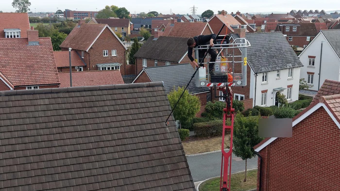 Operative in a raised access platform basket working a pole across a plain-tiled roof above a modern housing estate