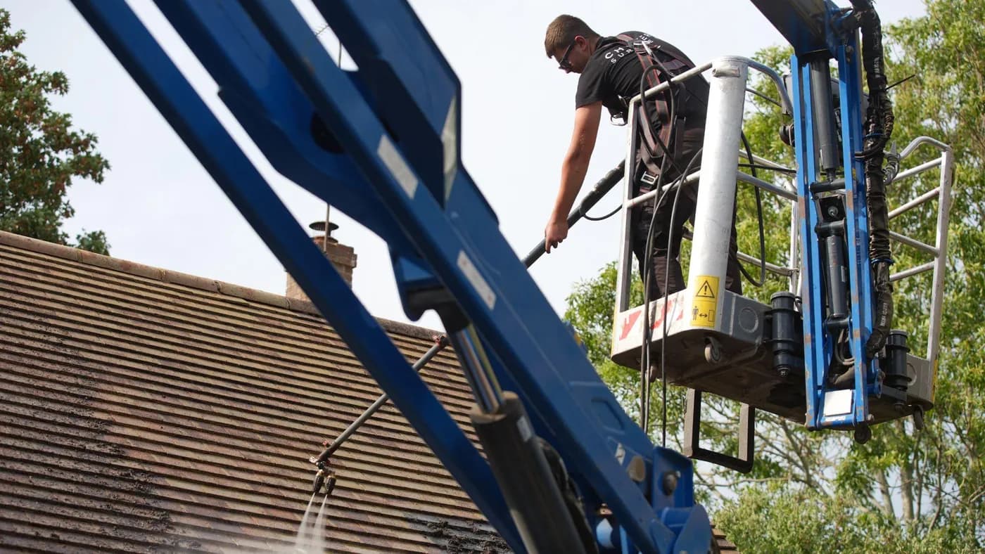 Operative in a harness on an access platform rinsing a clay tile roof with a twin-nozzle lance, trees behind