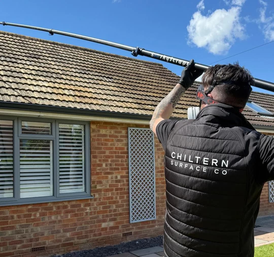 Operative standing in a garden raising a long carbon pole towards the eaves of a mossy concrete-tiled bungalow roof