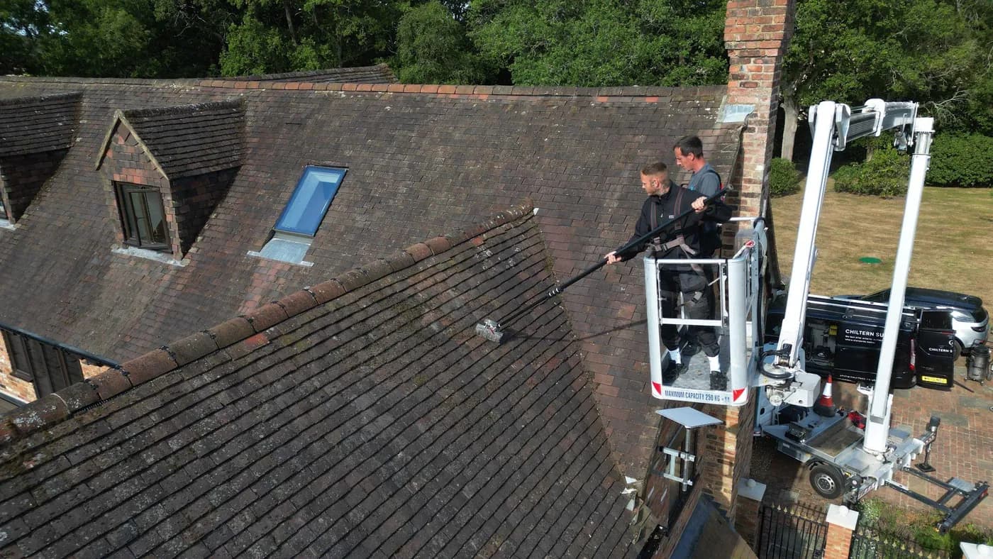 Two operatives in a raised platform basket brushing a weathered clay-tiled roof with a long pole, trees behind