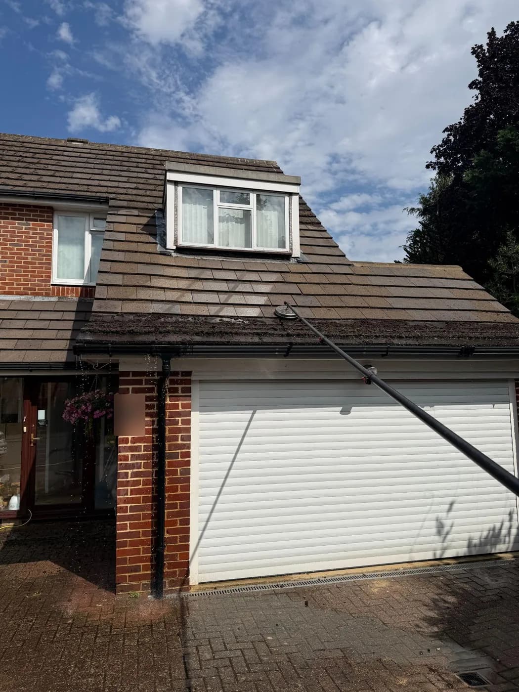 Pole-mounted rotary cleaner working the lower courses of a tiled roof over a garage, with a flat-roofed dormer above