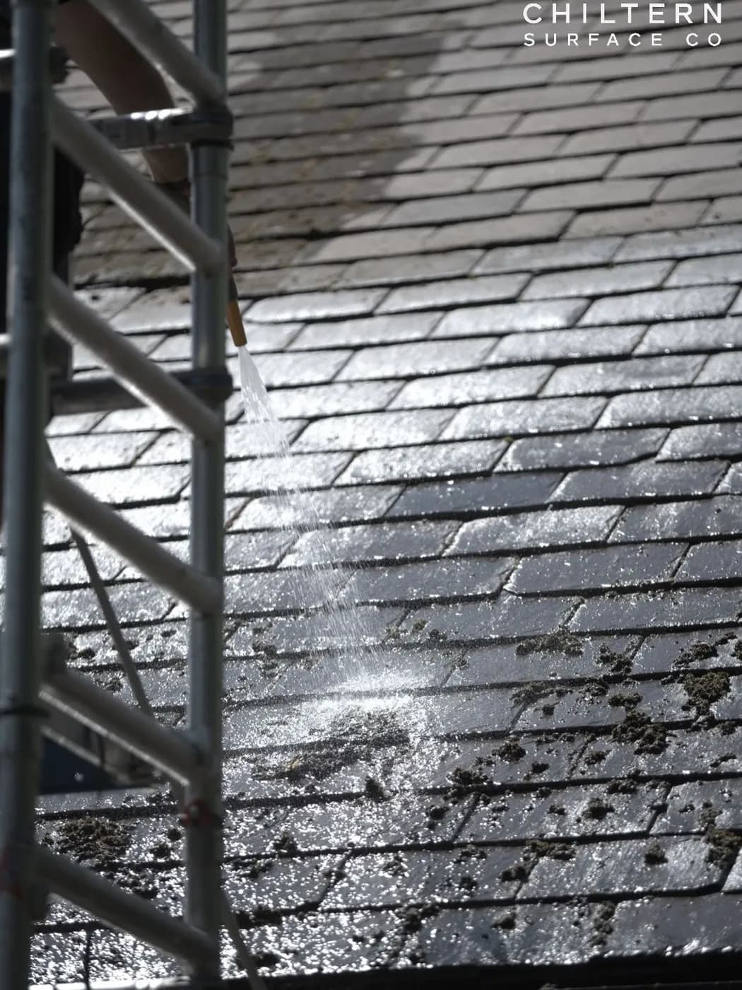 Operative on an access tower rinsing a slate roof at low pressure, loosened moss scattered across the lower slates