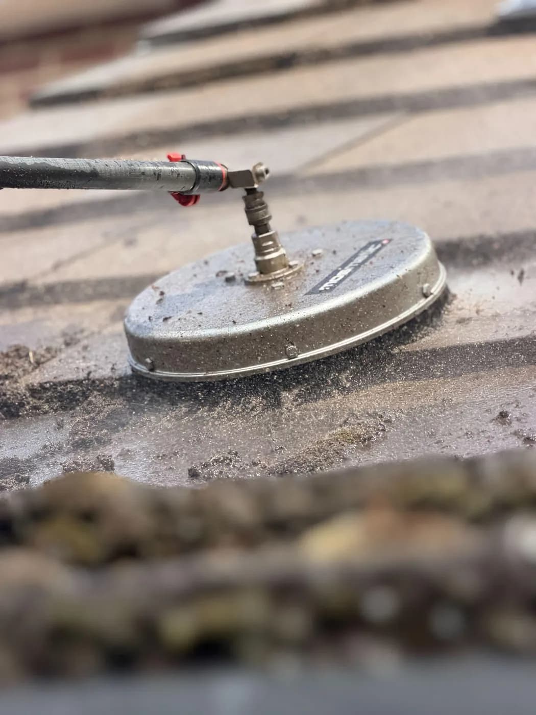 Close view of a pole-mounted rotary surface cleaner working across wet roof tiles, loosened moss scattered around it