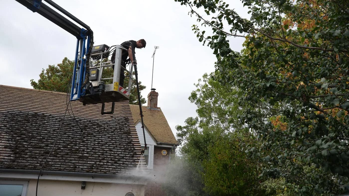 Operative on a boom lift platform rinsing a mossy tiled roof with a pole lance, mature trees beside the house