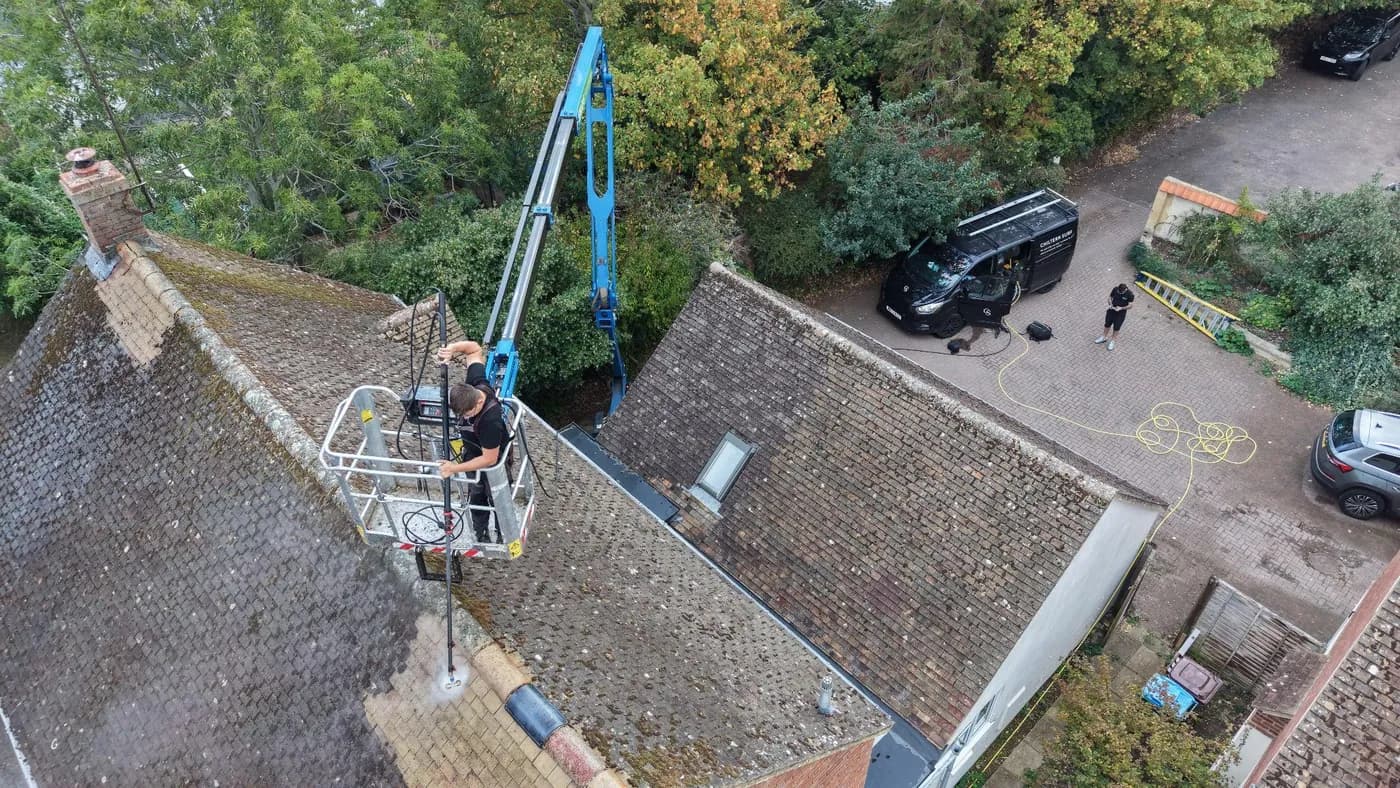 Operative in a boom-lift basket working a pole-mounted rotary cleaner across a mossy tiled roof, a cleaned strip behind