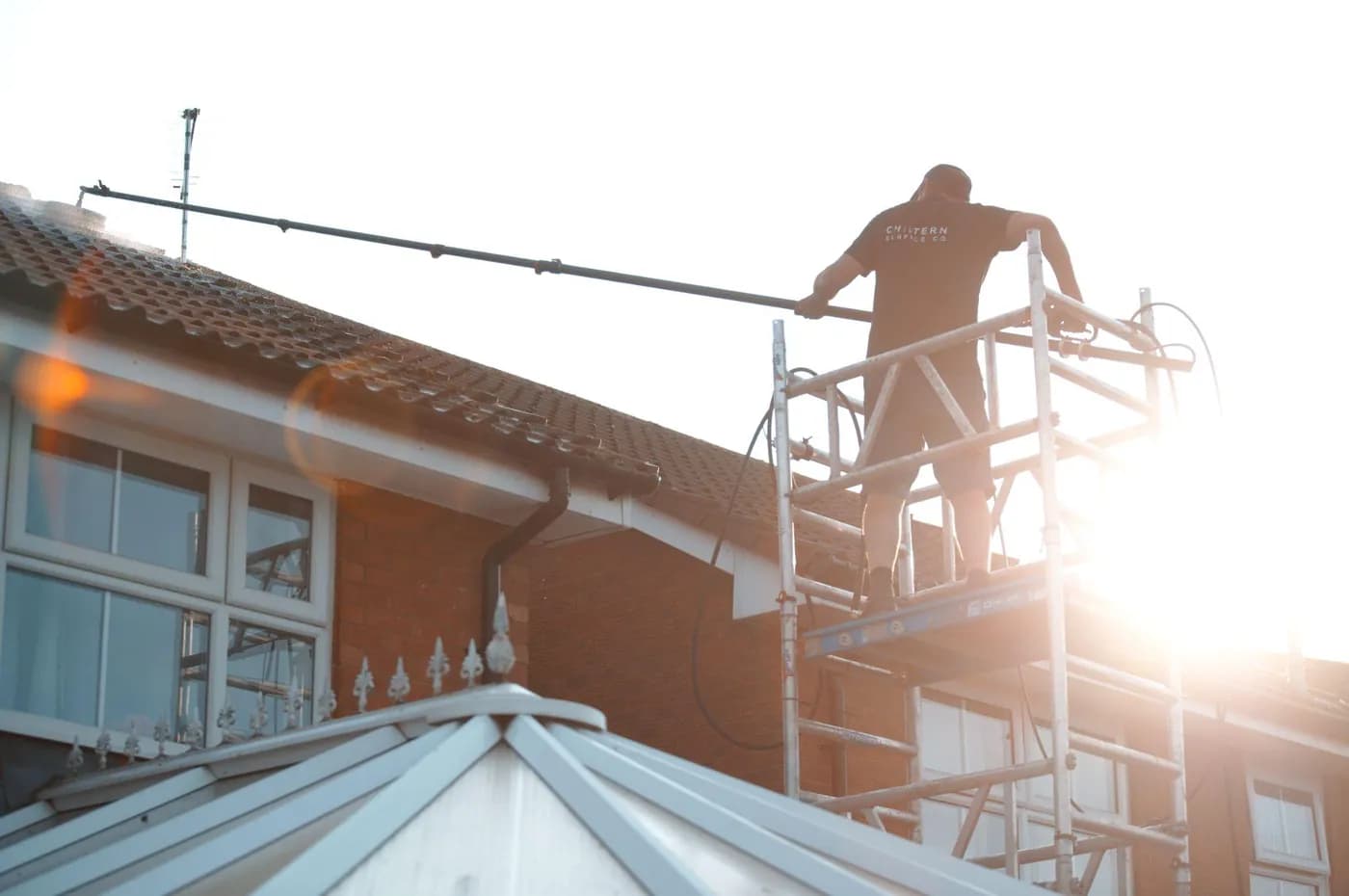 Operative on an access tower reaching across a tiled roof with a long pole, a conservatory roof in the foreground