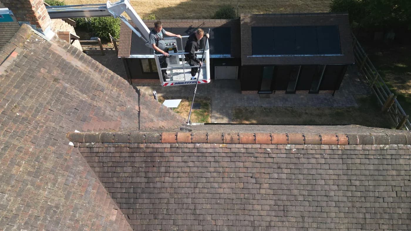 Two operatives in a raised platform basket brushing the ridge of a weathered clay plain-tile roof, seen from above