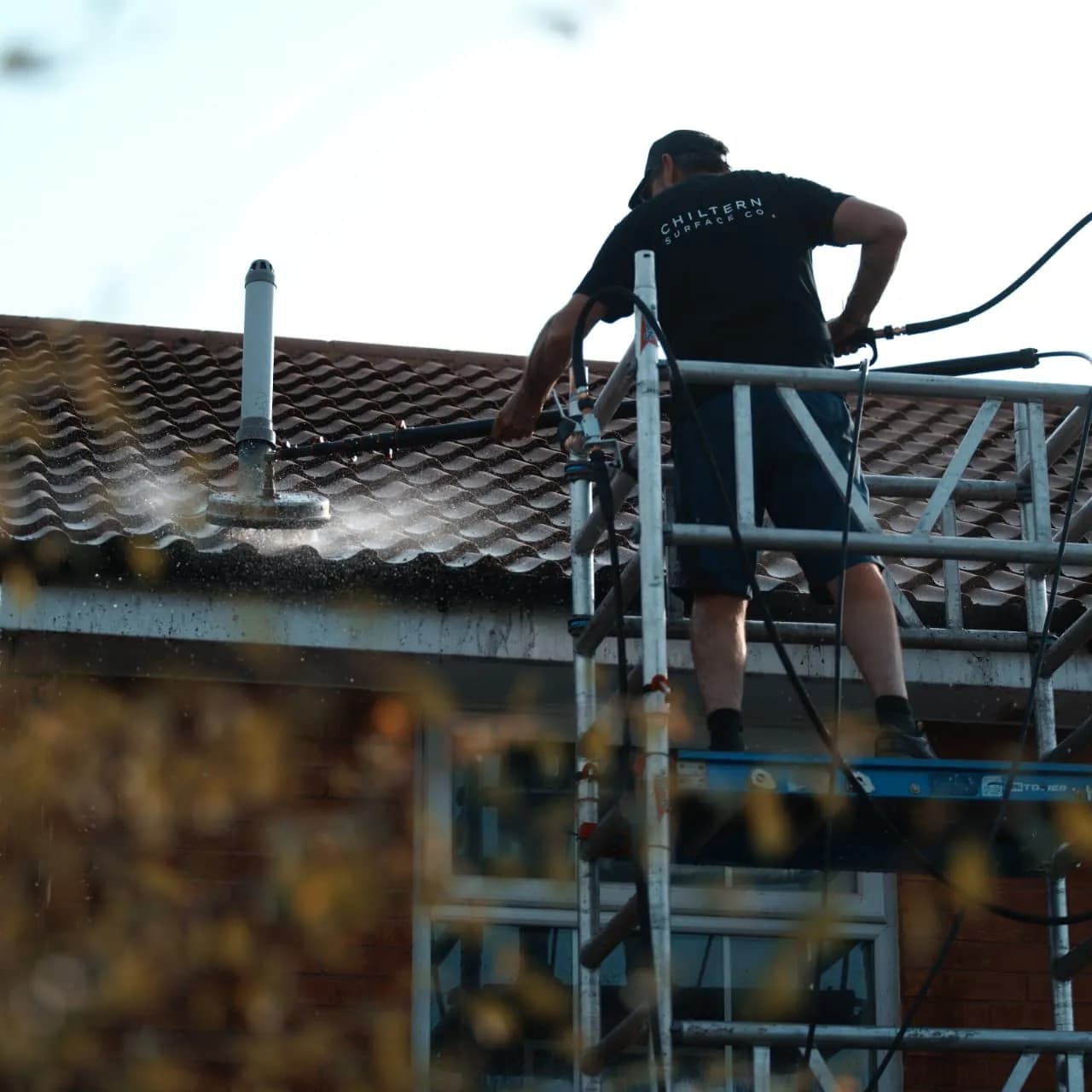 Operative on an aluminium access tower guiding a rotary surface cleaner across profiled roof tiles beside a flue