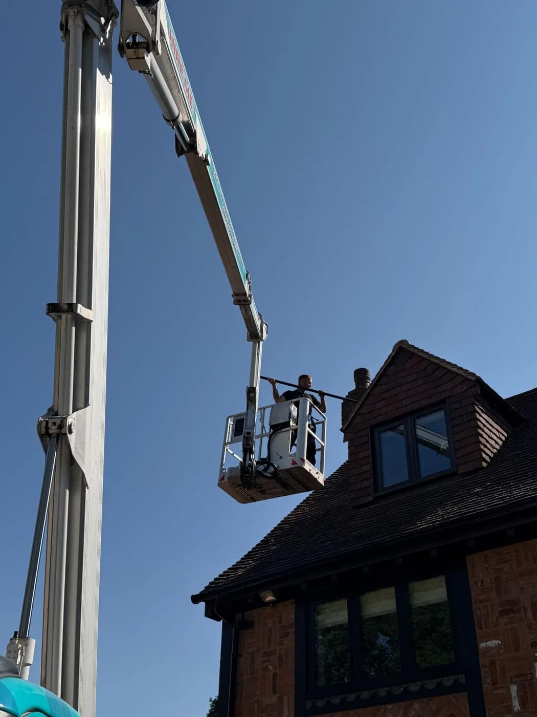 Operative in an elevated platform basket holding a long pole beside a clay-tiled roof with a brick dormer