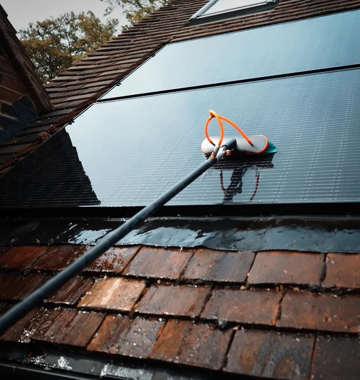 Water-fed brush head on wet black solar panels, a roof window above and wet clay tiles below