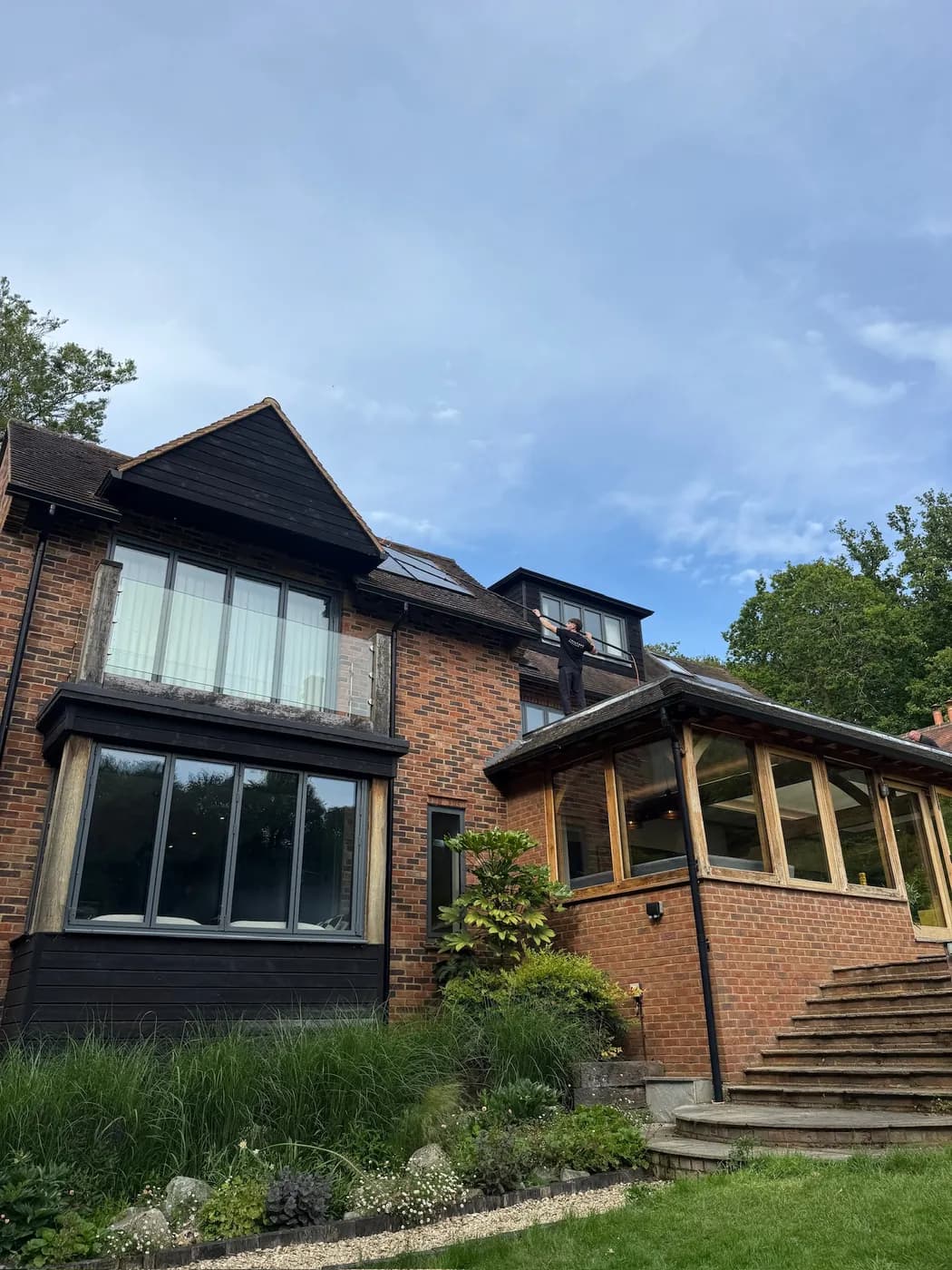 Wide view of a brick and timber house with an operative on a lower roof cleaning solar panels by pole