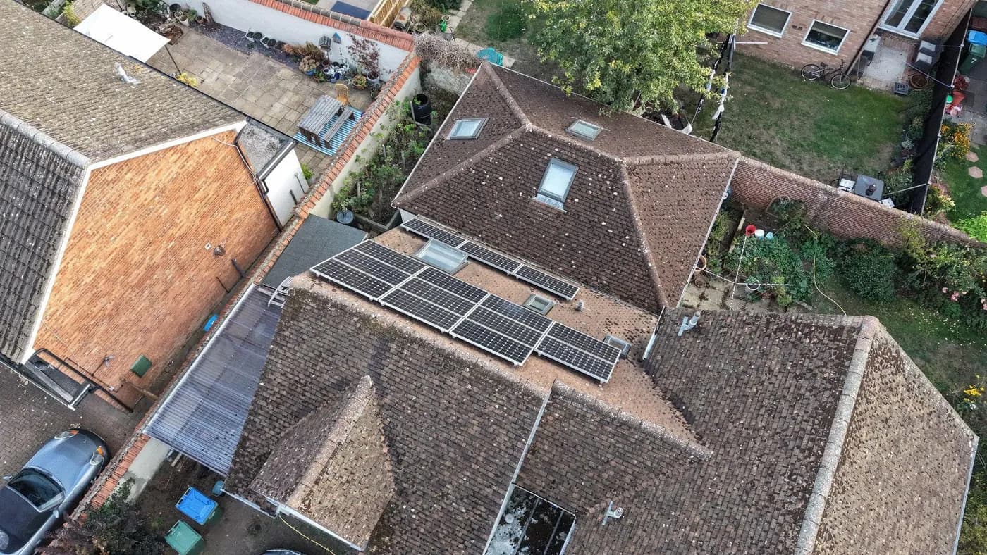 Drone view of a roof-mounted solar array set among weathered, mossy clay tiles, with roof windows and gardens below