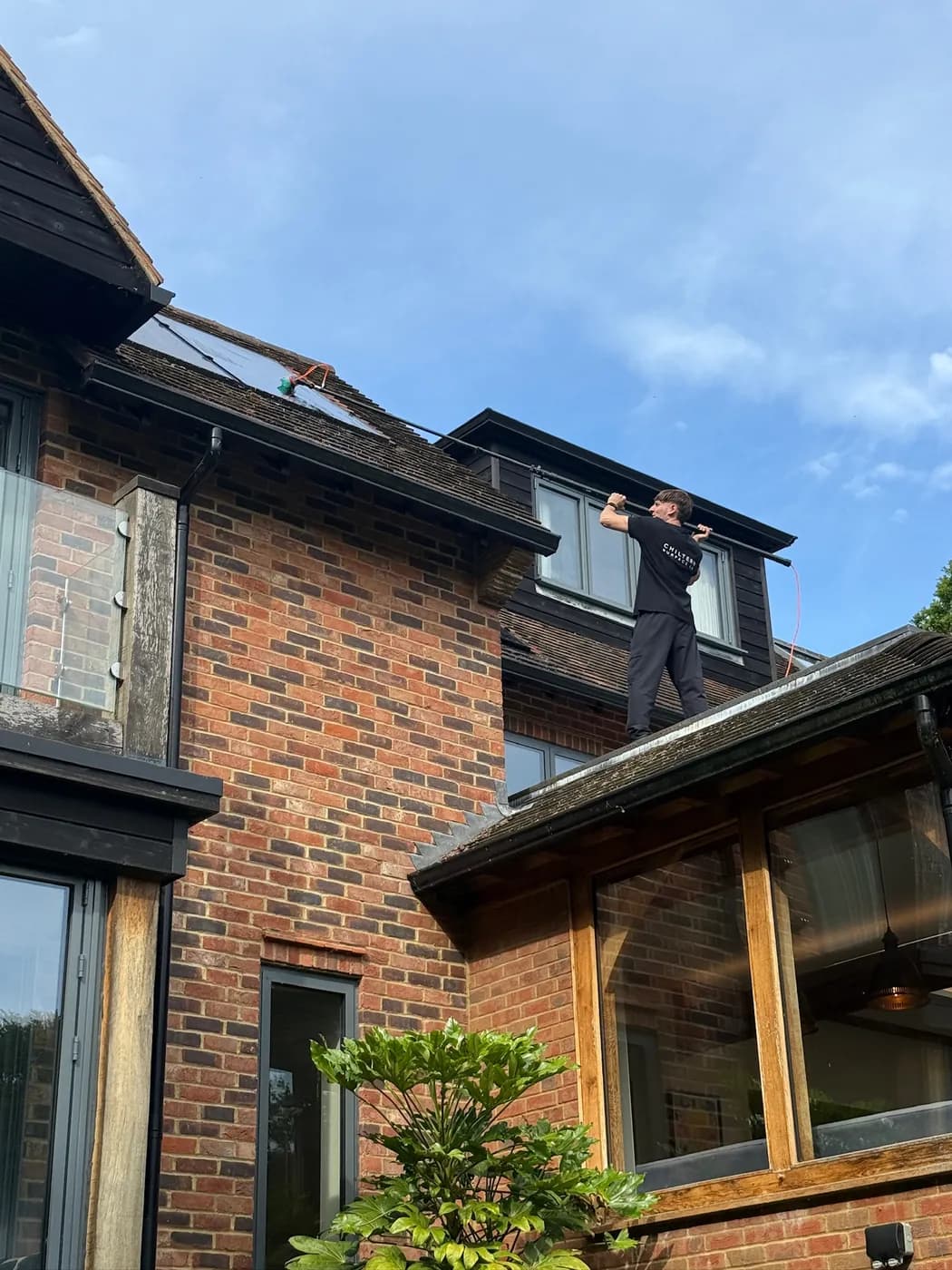 Operative on an extension roof edge guiding a pole brush over solar panels on a clay-tiled upper slope