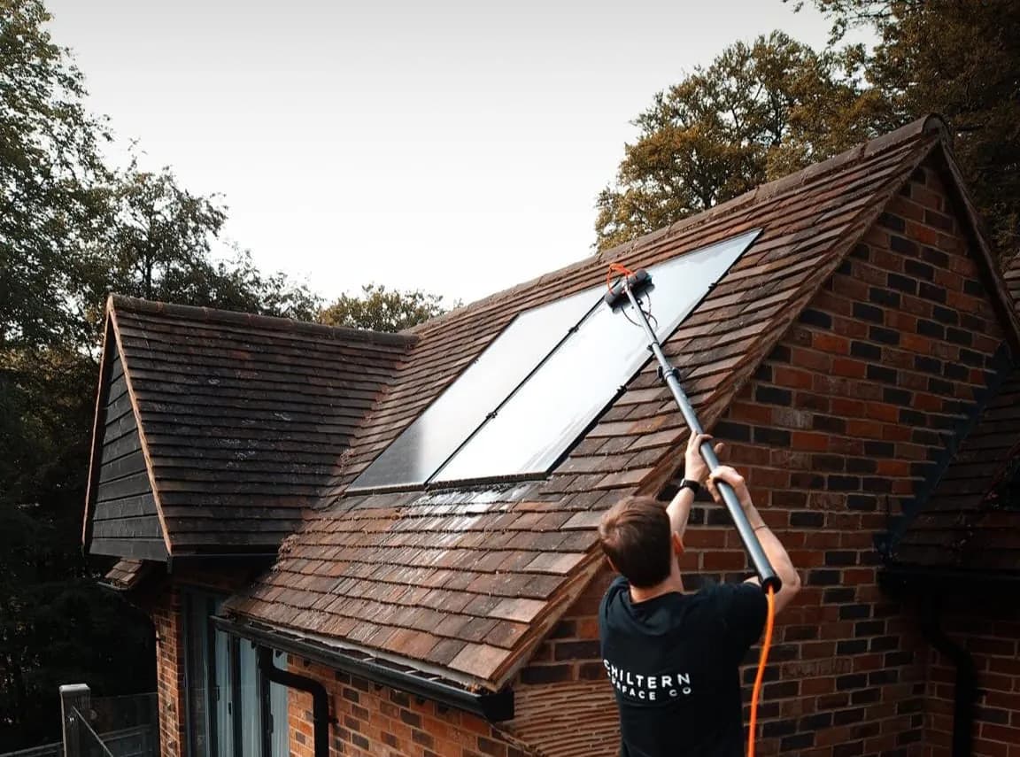 Operative reaching a long water-fed pole up to a pair of solar panels on a weathered clay-tiled roof