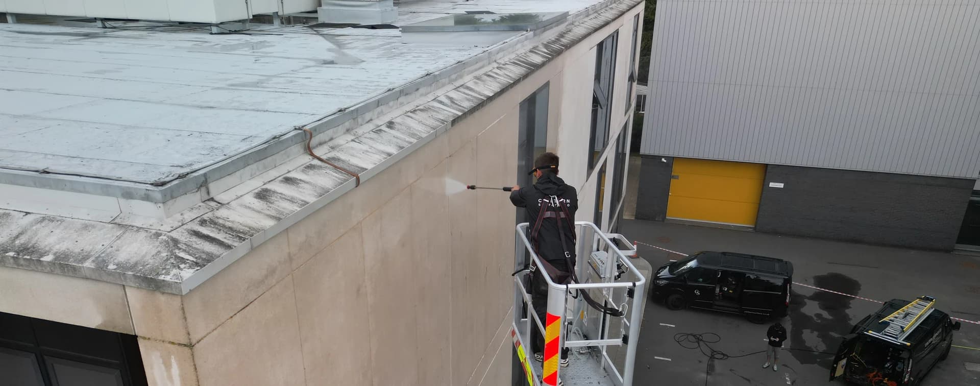 Aerial view from above of a technician washing a stained concrete elevation from a platform basket, vans and tape beyond