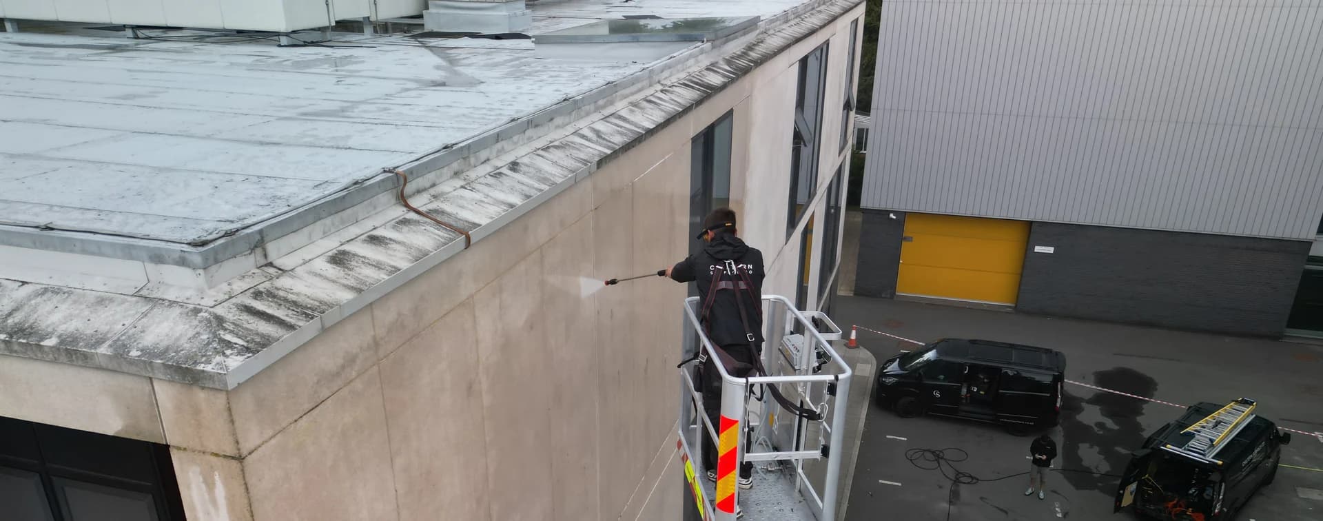 Aerial view from above of a technician washing a stained concrete elevation from a platform basket, vans and tape beyond