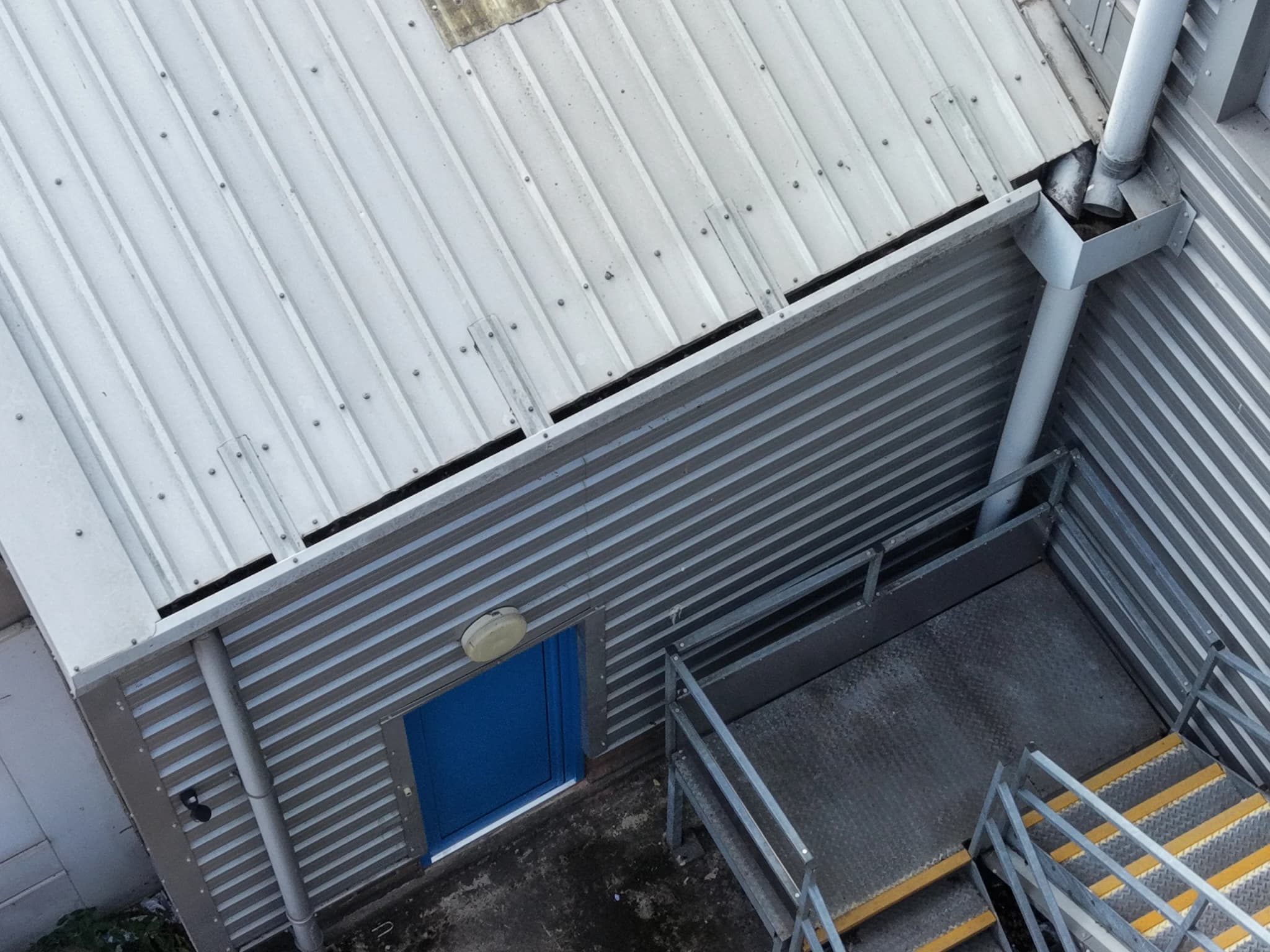Aerial view of a corrugated metal-clad wall with a blue door, a downpipe and a galvanised access stair