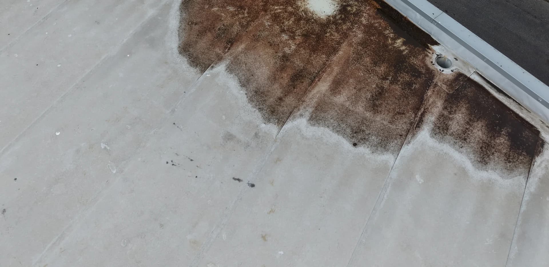 Aerial view of a flat membrane roof corner with a large rust-brown stain around an outlet beside the parapet