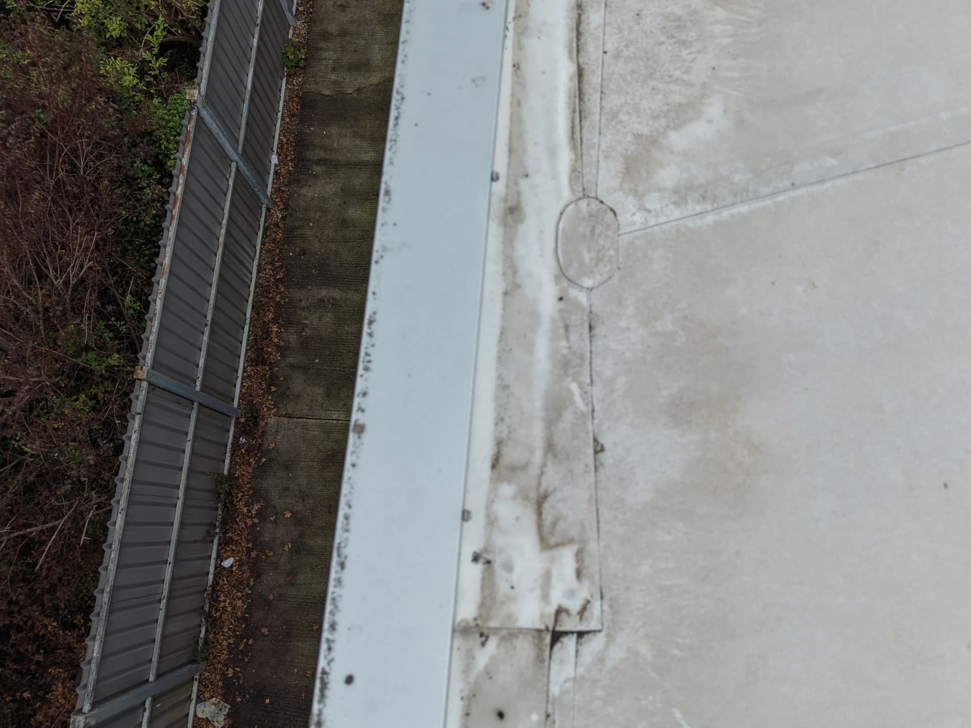 Aerial view of a flat roof edge and parapet beside a fence and trees, with light staining