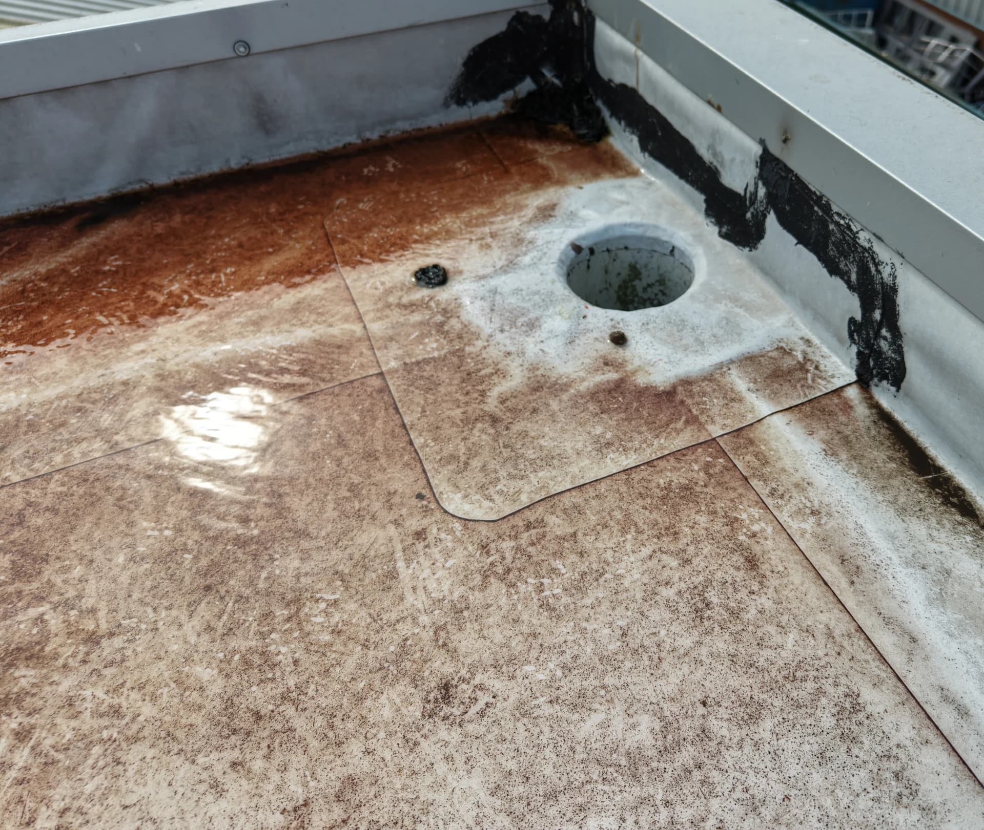 Aerial view of a roof outlet and a cracked tar repair at a parapet corner with rust staining on the membrane