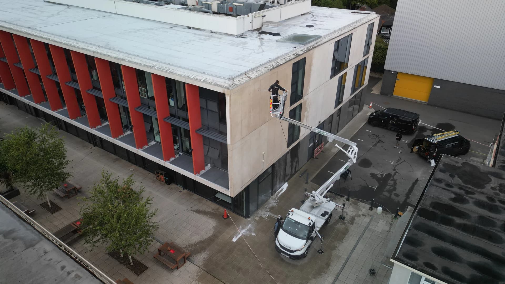 Aerial view of a technician in the basket of a truck-mounted access platform washing a concrete-panelled building, vans and tape beyond