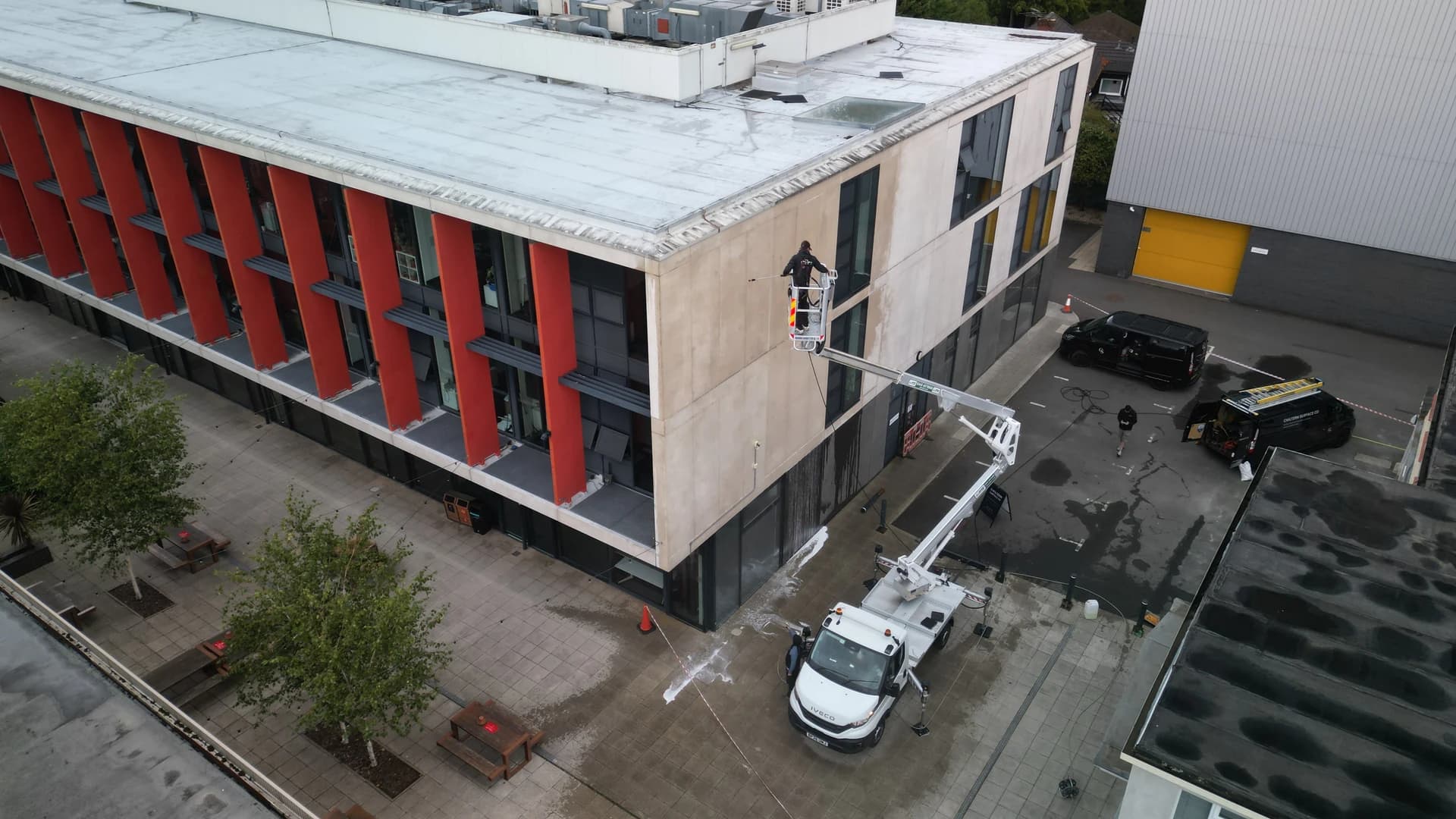 Aerial view of a technician in the basket of a truck-mounted access platform washing a concrete-panelled building, vans and tape beyond