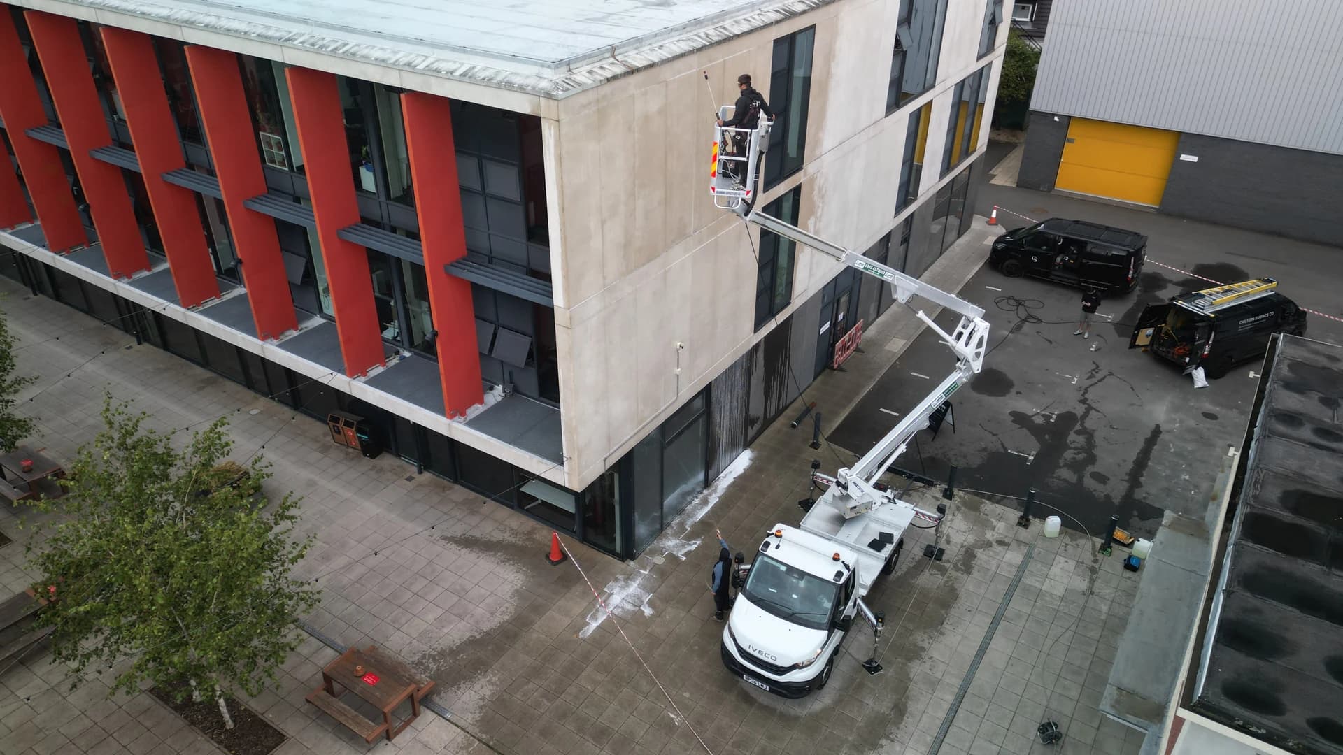 Aerial view of a truck-mounted access platform with a technician in the basket cleaning a concrete-panelled elevation, vans beyond