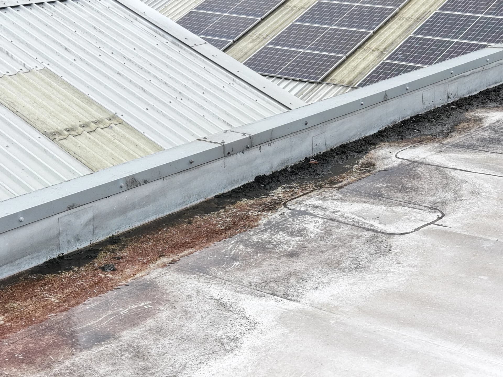 Aerial view of debris and silt in a valley gutter against a parapet, a patched seam and rooftop panels behind