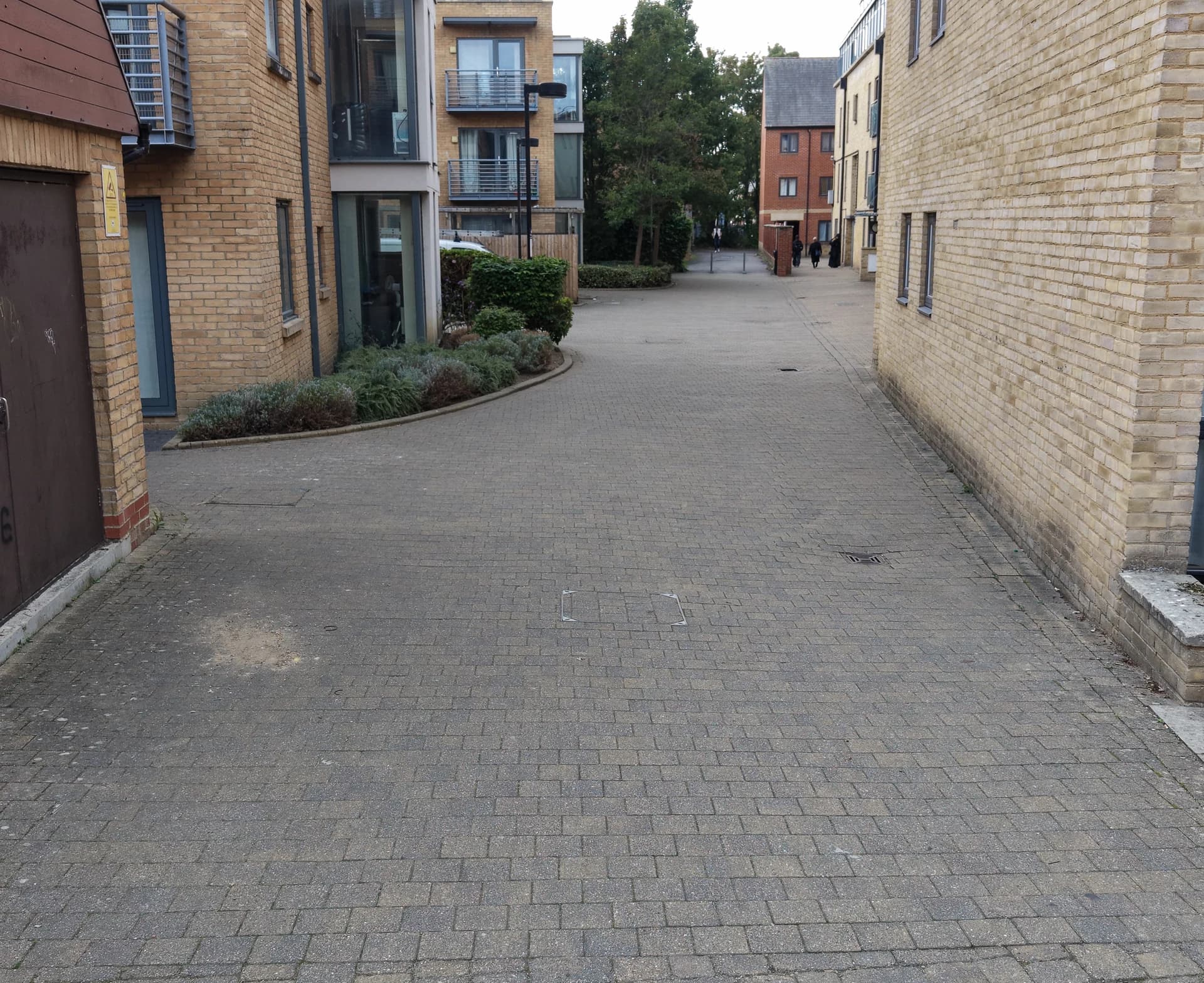 Block-paved walkway between brick buildings, with planting at the side and a few distant pedestrians