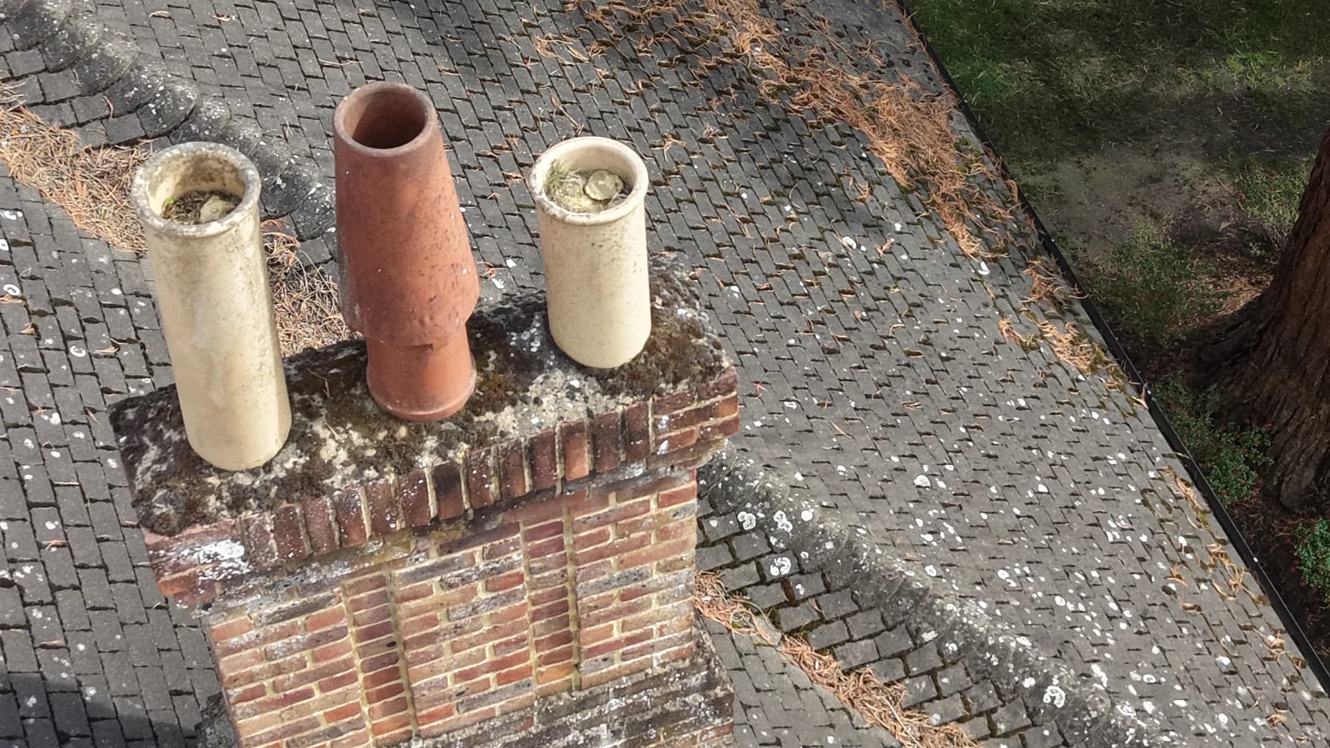 Chimney stack with three pots rising from a tiled roof slope spotted with moss and lichen
