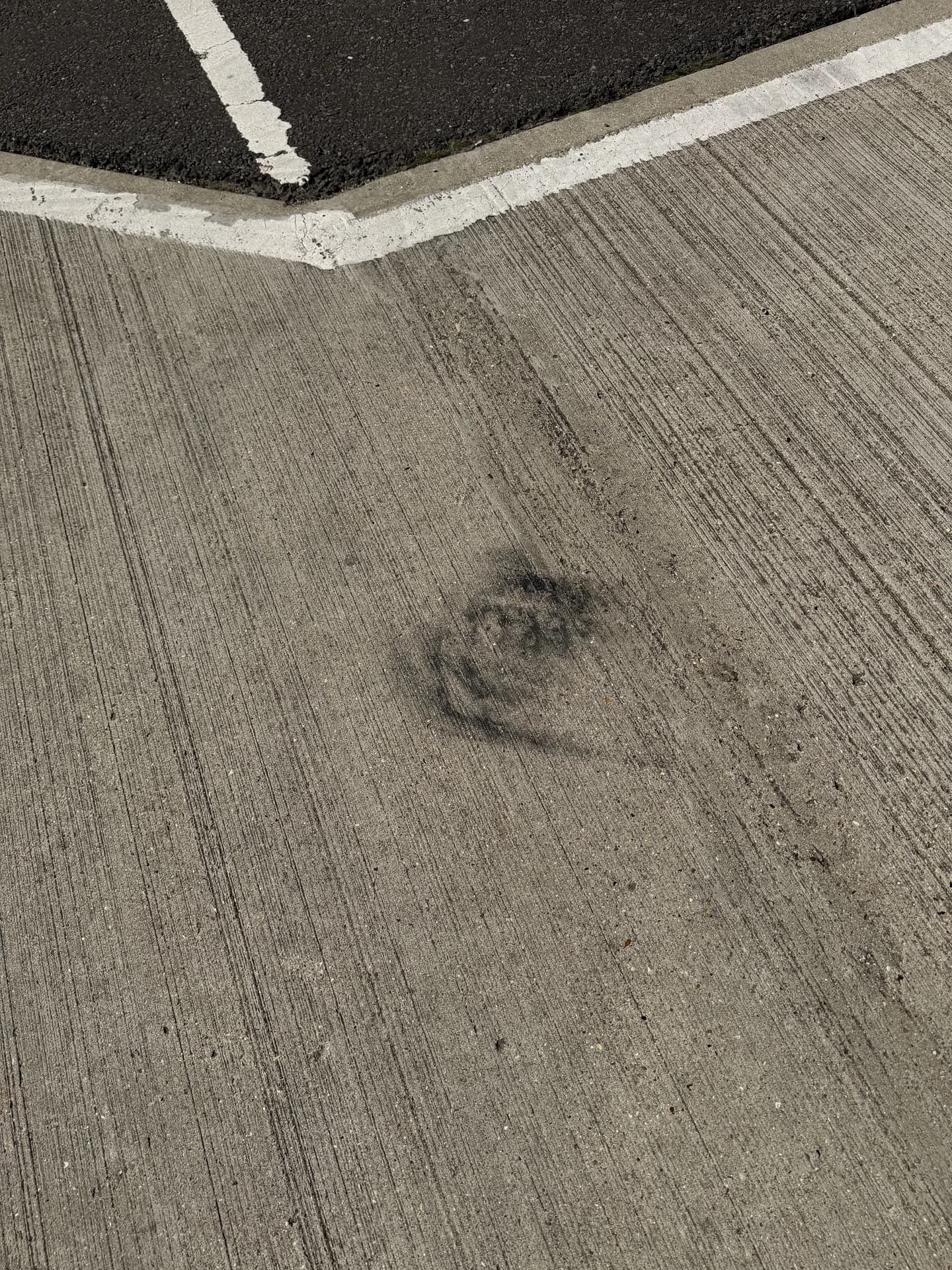 Circular tyre and oil mark on a car-park surface beside a white bay line and a tarmac edge