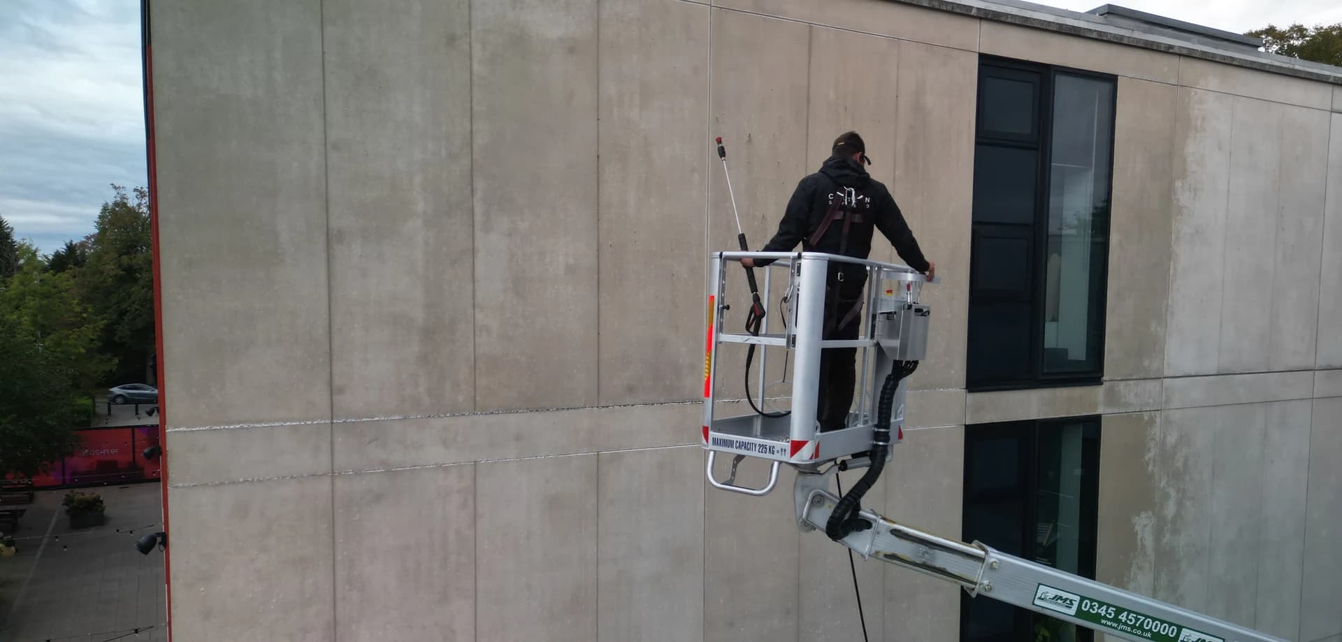 Close aerial view of a technician from behind, harnessed in a platform basket, washing a concrete facade panel