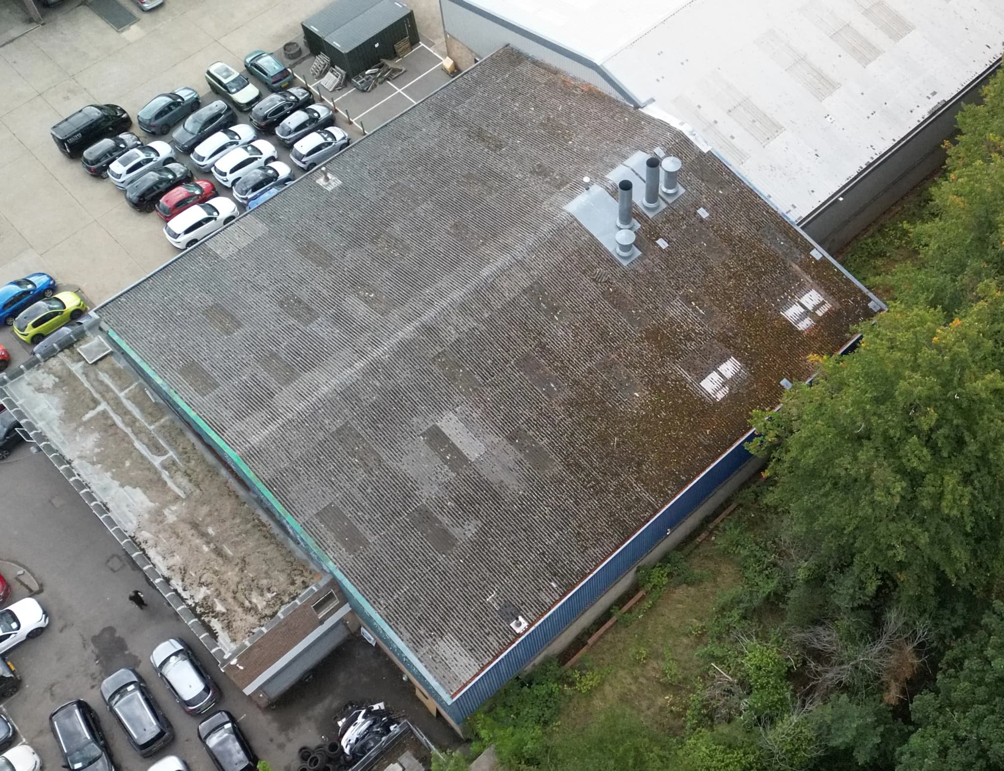 High oblique aerial view over a whole fibre-cement industrial roof with vents, a car park to one side and trees beyond