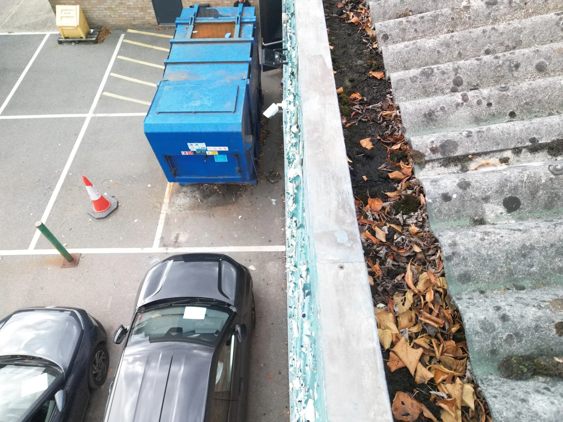 Looking down into a leaf-choked gutter beside the roof, blue skips and cars in the yard below.