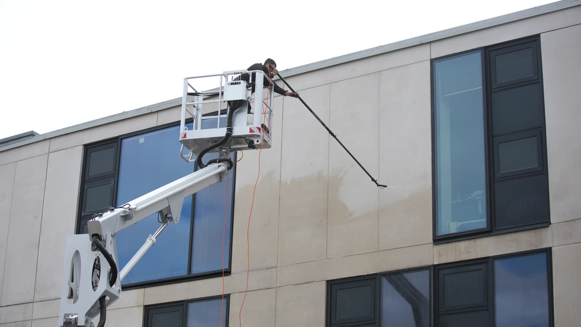 Low-angle view of a technician in a truck-mounted platform basket washing a stone-coloured panel, a clean line against the dirtier panel