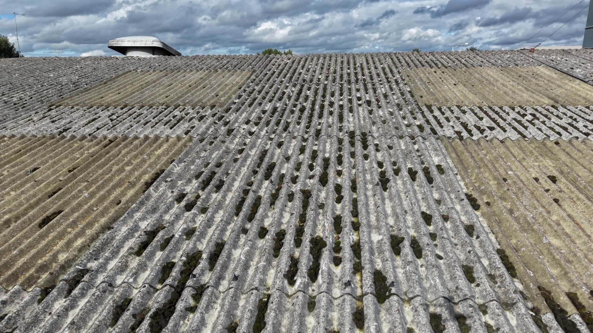 Low oblique aerial view along a mossed fibre-cement roof slope to the ridge, with a roof vent