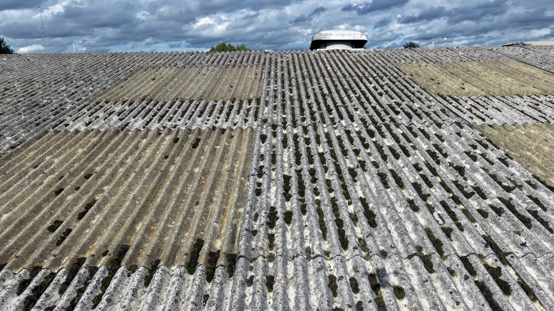 Low oblique view along a moss-covered fibre-cement roof slope rising to the ridge under heavy cloud