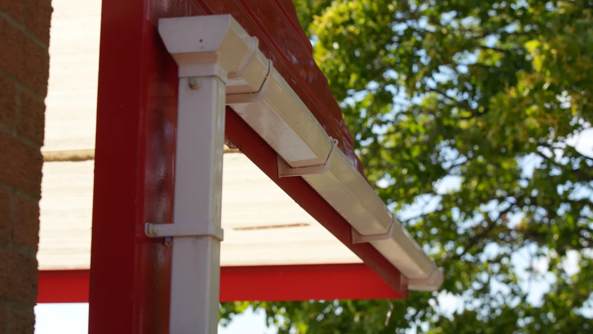 Red steel canopy post with a rectangular white downpipe and gutter; bright tree behind.