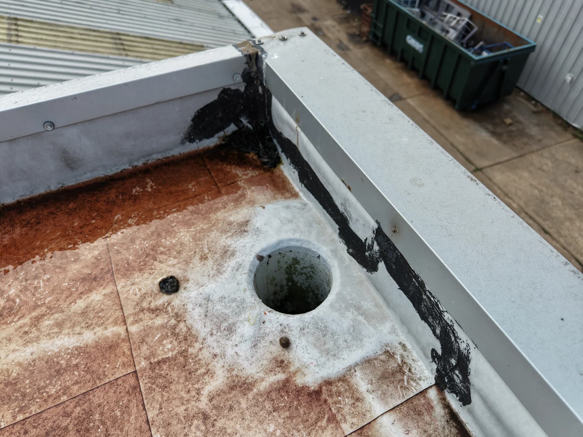 Same outlet and tar-repaired parapet corner, seen with a green skip behind.