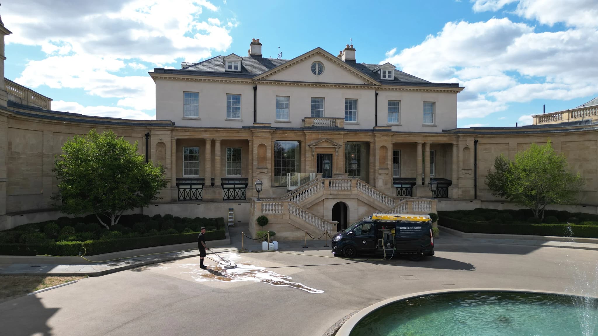 Technician side-on with a surface cleaner on a foamed patch of paving, a van and stanchion cordon in front of a stone-built hotel