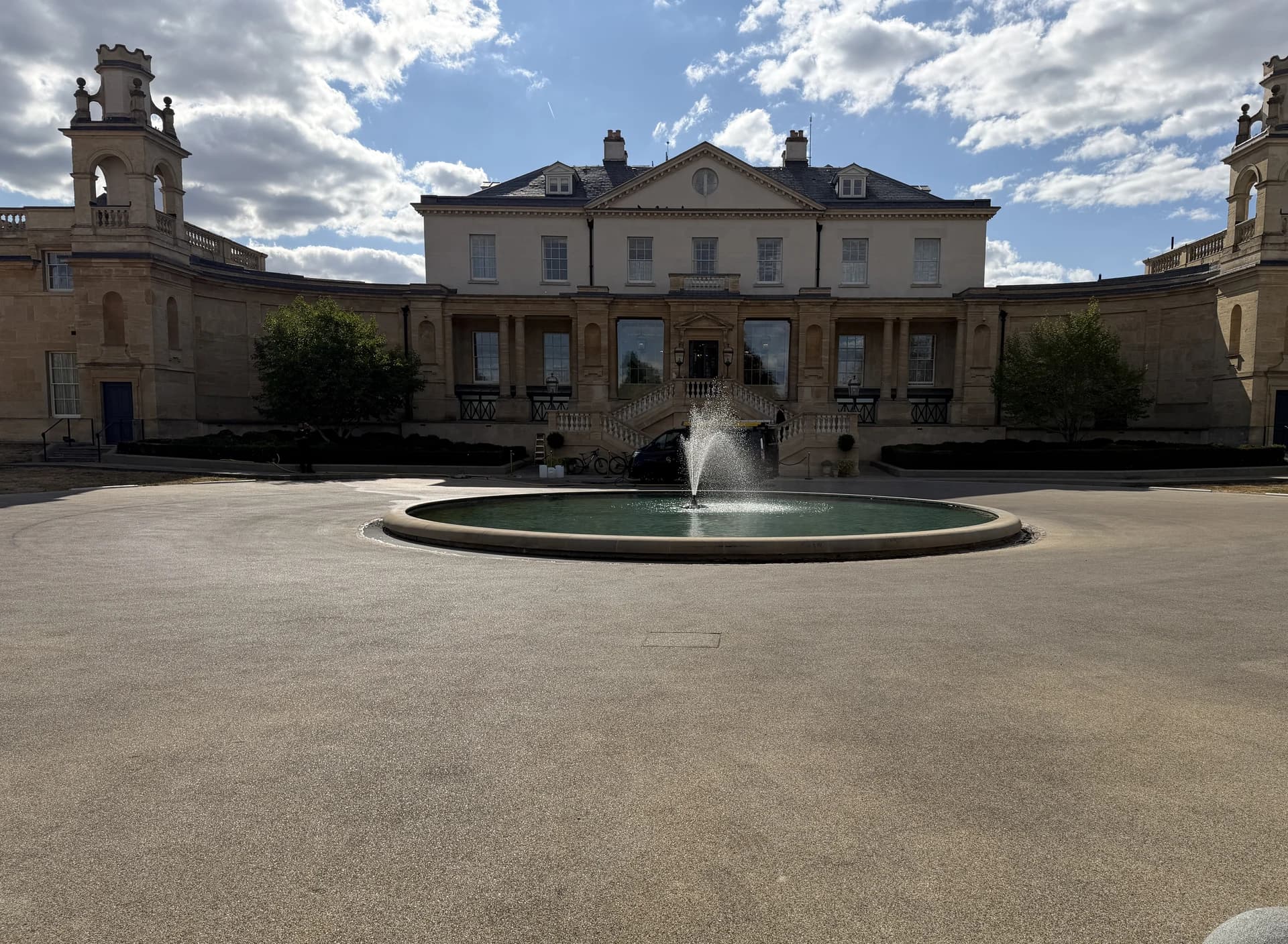 Wide empty paved forecourt with a fountain in strong sunlight and a stone-built building behind it