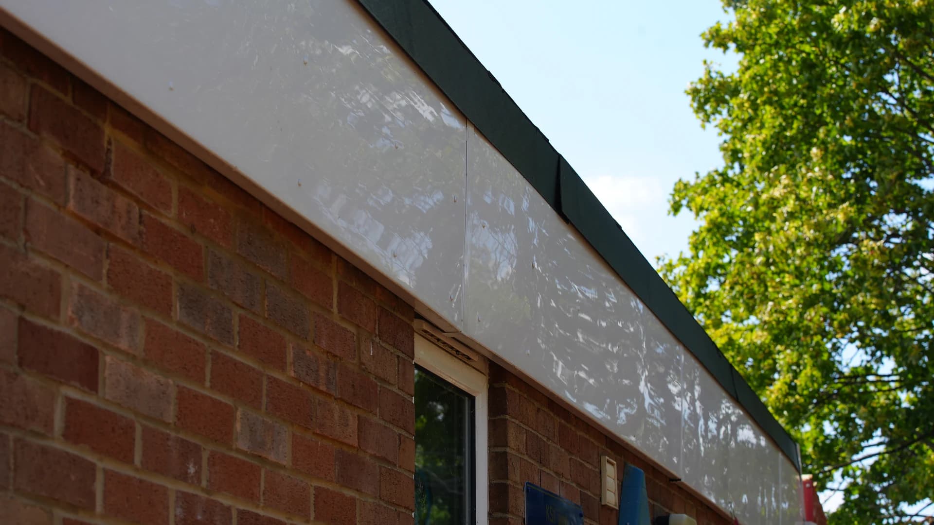 Wide upward view of a clean white fascia panel along a brick wall under a flat roof, trees at right.